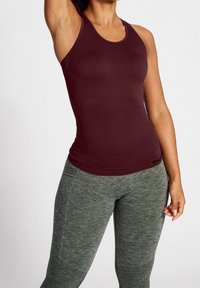 Donna che indossa un top aderente color marsala e leggings grigi con texture, posando con un braccio alzato davanti a uno sfondo semplice.