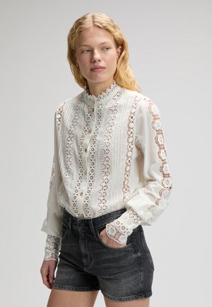 Młoda kobieta o falowanych blond włosach ubrana w białą koronkową bluzkę w kwiatowe wzory i czarne krótkie spodenki jeansowe, z ręką w kieszeni, na szarym tle.