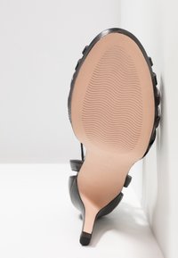 Sandale à talon haut noire reposant sur le côté, montrant une semelle avec un motif ondulé et un embout de talon fin.
