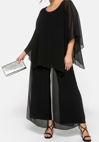 Černý průhledný top s kulatým výstřihem zdobeným korálky, doplněný širokými kalhotami. Doplňuje ho stříbrná kabelka a černé sandály na pásky.
