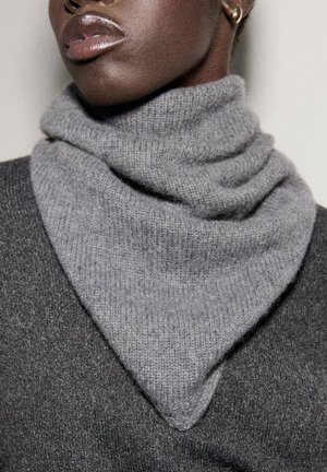 Bufanda gris de punto con forma triangular, textura suave y capas de pliegues. Lleva puesta sobre un suéter oscuro, mostrando una rica paleta de colores apagados.