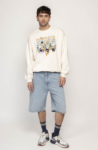 Hanorac crem cu un imprimeu grafic cu alergători, asortat cu pantaloni șort din denim ușor albastru. Se evidențiază șosete cu dungi navy și adidași multicolori.