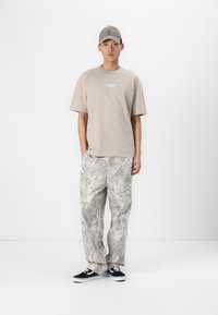 Beige Baumwoll-T-Shirt mit weißem Logo, hellgraue gemusterte Hose, schwarze Sneaker mit weißen Sohlen und passende graue Kappe.