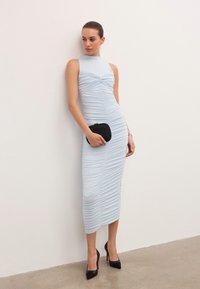 Ženska v svetlo modri, nabrani brezrokavni midi obleki drži črno torbico, nosi črne visoke pete in stoji ob beli steni.