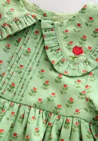 Grünes Kleid mit floralem Muster aus roten Blumen und grünen Blättern. Rüschensaum mit gesticktem Rosendetail und gerafftem Taillenbereich.