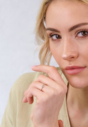 Zarter silberner Ring mit einem zentralen Stein, getragen an einer Hand mit neutralem Maniküre; der Hintergrund zeigt sanfte, gedämpfte Farben und natürliche Texturen.