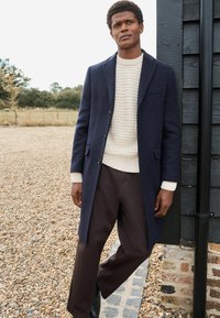 Abrigo de lana navy con un diseño entallado, suéter de punto crema y pantalones marrones. Presenta un cierre de un solo boton y una textura sutil.