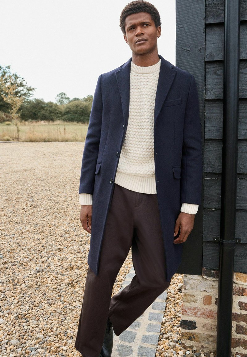 Abrigo de lana navy con un diseño entallado, suéter de punto crema y pantalones marrones. Presenta un cierre de un solo boton y una textura sutil.