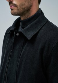 Crna vuna jakna sa čvrstim ovratnikom, zatvaranjem na zip i teksturiranom tkaninom. Nosena preko tamnog turtleneck pulovera, prikazuje prilagođeni siluetu.