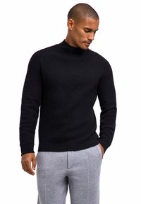 Schwarzer Strickpullover mit hohem Kragen, geripptem Muster und langen Ärmeln, kombiniert mit grauer Jogginghose. Der Schnitt ist locker und modern.