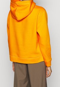Sudadera con capucha naranja de corte relax, fabricada en un tejido texturizado y con costuras visibles. Combinada con pantalones marrones holgados.