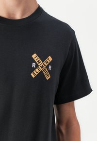 Černé bavlněné tričko s grafikou, která na hrudi zobrazuje béžový a bílý text ve tvaru X s nápisem "TIMBER! ELEMENT!".