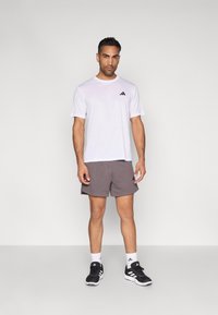 Camiseta desportiva branca de mangas curtas com logótipo preto, combinada com calções cinzentos e sapatilhas desportivas pretas com detalhes brancos.