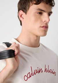 Cream t-shirt with red embroidered "calvin klein" text, smooth texture, round neckline, paired with a black bag handle held in the right hand.