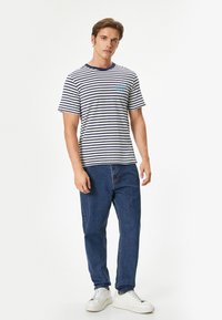 Camiseta de manga corta a rayas en azul marino y blanco con cuello redondo. Combinada con jeans azules y zapatillas blancas. Detalle del logo visible en el pecho.