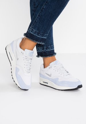 Osoba nosząca białe i jasnoniebieskie buty Nike z widocznymi poduszkami powietrznymi oraz ciemnoniebieskie podwinięte dżinsy na białym tle.