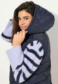 Gilet trapuntato blu navy con un ampio cappuccio e maglione in maglia viola chiaro con strisce blu scuro. Il tessuto ha una texture liscia e lucida.