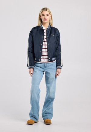 Vrouw staand, gekleed in een marineblauw bomberjack met witte strepen, een gestreept poloshirt, lichtblauwe jeans en lichtbruine suède schoenen, tegen een grijze achtergrond.