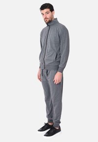 Giacca sportiva grigia con zip e colletto alto, dotata di tasche laterali, abbinata a pantaloni jogger coordinati e sneaker nere.