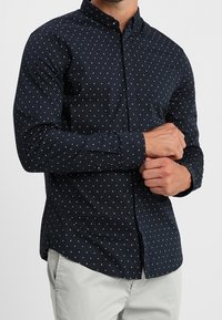 Námořnická modrá košile na knoflíky s malým bílým geometrickým vzorem, dlouhé rukávy a špičatý límec. Tkanina vypadá hladce a lehce.