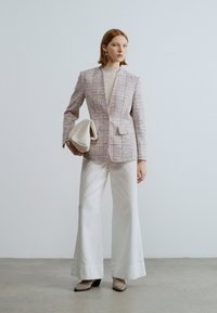Veste en tweed rose et blanc sur mesure avec un revers à encoches, associée à un pantalon blanc à jambes larges et une pochette crème. Le modèle porte des talons noirs.
