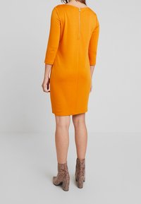 Orange, fitted dress with three-quarter sleeves, features a back zipper. Paired with textured, snake-print ankle boots on a neutral background.