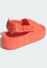 Coral red sandals with a textured surface, adjustable straps, and thick, cushioned soles. Features subtle logo detailing on the side.