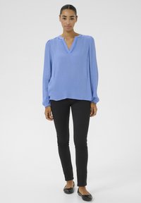Plava bluza dugih rukava s V-izrezom i teksturiranim materijalom; uparena s crnim slim-fit hlačama i crnim baletankama.