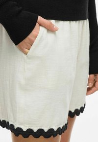 White shorts with a textured fabric, featuring black scalloped trim at the hem and a side pocket, paired with a black knitted sweater.