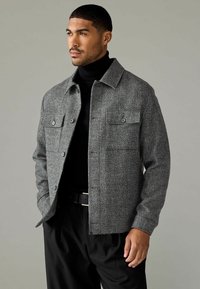 Jachetă texturată gri cu un design cu guler, două buzunare pe piept, închidere cu butoni și o croială lejeră, asortată cu un pulover negru cu guler înalt.