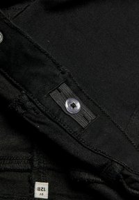 Gros plan sur la ceinture d’un vêtement noir montrant un élastique réglable avec bouton et une étiquette de taille en tissu indiquant 128.