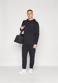 Sort hættetrøje og sweatpants sæt i blødt stof, lomme detaljer på hættetrøjen, parret med hvide sneakers og en sort duffel taske.