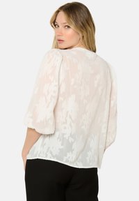 Blouse blanche et légère avec texture florale, manches bouffantes et coupe ample. La vue de dos montre des détails subtils et un ourlet arrondi.