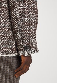 Manche de veste texturée marron et blanche avec bouton noir et bord effiloché, associée à un pantalon marron à motif, main reposant sur le côté.