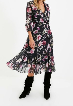 Vrouw draagt een zwarte bloemen-midi-jurk met ruches en driekwart mouwen, gecombineerd met zwarte slappe kniehoge laarzen.