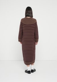 Μακρύ, καφέ πλεκτό φόρεμα με σκούρες μοβ οριζόντιες ρίγες. Διαθέτει στρογγυλή λαιμόκοψη και μακριά μανίκια, συνδυασμένο με μαύρα παπούτσια και λευκές κάλτσες.