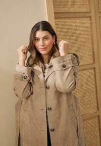 Femme ajustant le col d'un trench-coat beige, debout à l'intérieur devant un mur clair et un panneau en bois tissé en arrière-plan.