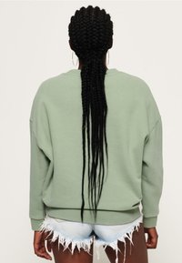 Jasnozielona bluza z luźnym krojem i opuszczonymi ramionami, zestawiona z denimowymi szortami z przetarciami. Długie, splecione w warkocz włosy opadają na plecy.