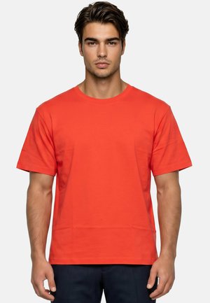 Jonge man met bruin haar die een effen rood T-shirt met korte mouwen en een donkere broek draagt en recht vooruit kijkt tegen een neutrale achtergrond.