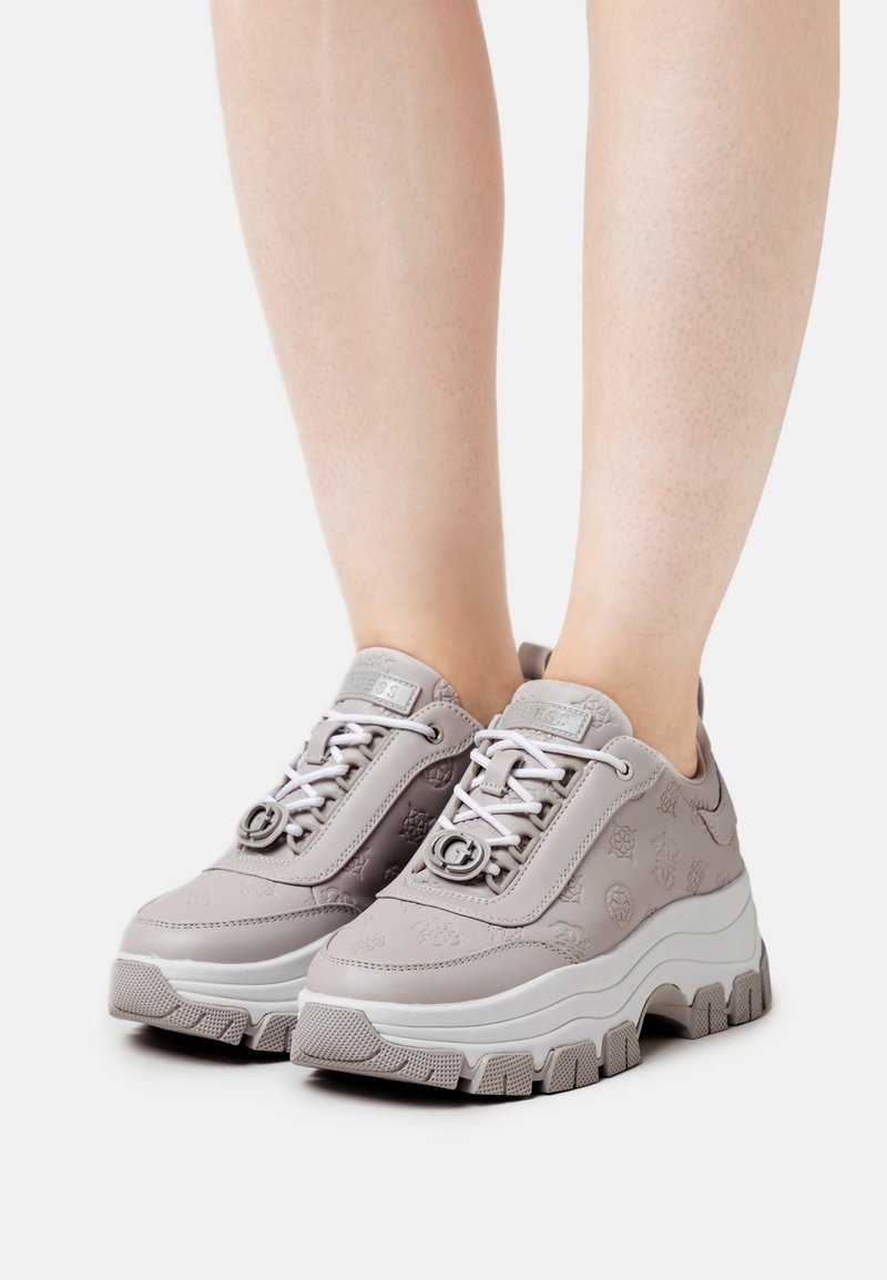 Sneaker sintetiche grigie con dettagli testurizzati, lacci bianchi e suola bianca spessa. Presentano una punta arrotondata e accenti del logo sulla linguetta.