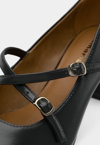 Μαύρο δερμάτινο παπούτσι με στρογγυλή μύτη, που διαθέτει σταυρωτή λογόθρα και μεταλλικές πόρπες. Ο εσωτερικός χώρος είναι καφετής με λεία υφή.