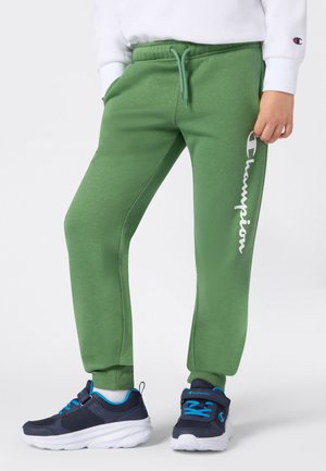 Kind dat groene Champion-trainingsbroek draagt en marineblauwe sneakers met blauwe accenten, terwijl hij met zijn handen in de zakken staat.