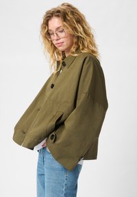Olive rohelises värvitoonis lühike jakk, mis on valmistatud siledast kangast, nuppude sulgemisega, suurte varrukatega ja küljetaskutega.