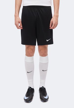 Férfi fekete Nike focishortban, fehér térdig érő Nike zokniban és fekete Nike stoplis cipőben világoskék részletekkel, kezei lazán vannak.