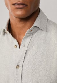 Camisa en espiga de color gris claro con cuello abotonado, que presenta botones beige y una tela suave y texturizada. Los detalles incluyen una sutil abertura en el cuello.