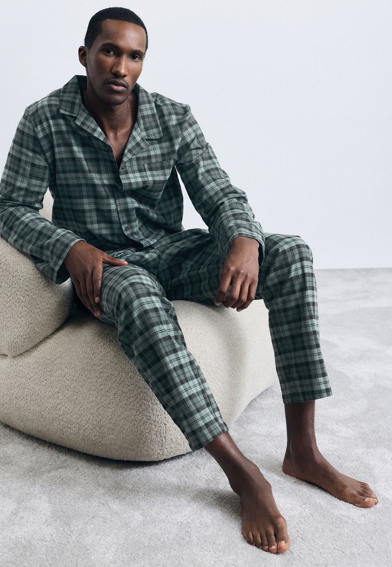 Πράσινο και γκρι καρό σετ πιτζάμας φτιαγμένο από μαλακό ύφασμα με κολάρο και κομψά παντελόνια, φορεμένο από ένα καθιστό μοντέλο σε μια υφασματική επιφάνεια.