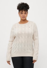 Pull en tricot torsadé beige clair avec un col rond, des poignets et un ourlet côtelés. Présente un petit détail de logo sur la poitrine.