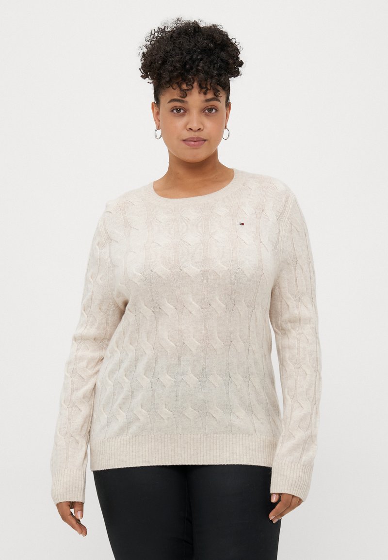 Pull en tricot torsadé beige clair avec un col rond, des poignets et un ourlet côtelés. Présente un petit détail de logo sur la poitrine.