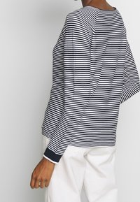 Chemise à manches longues rayée bleu marine et blanche, avec un col rond, des fentes latérales et des poignets côtelés, associée à un pantalon blanc.