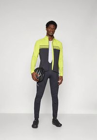 Ποδηλατική στολή με φωτεινό κίτρινο μπουφάν και μαύρα κολάν, με αντανακλαστικές ρίγες και διχτυωτό ένθετο για αερισμό. Κρατείται μαύρο κράνος.